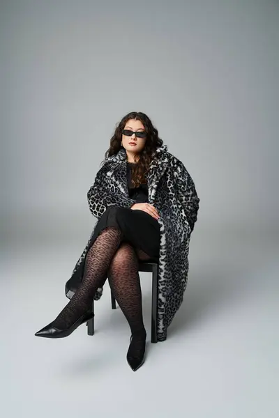 Verführerische Plus-Size-Frau in Leopardenfell-Mantel und Sonnenbrille auf schwarzem Stuhl mit überkreuzten Beinen sitzend — Stockfoto