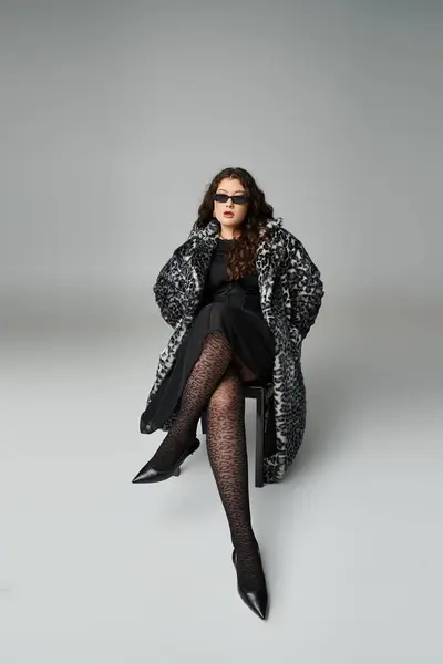 Belle femme courbée en manteau de fourrure de léopard et lunettes de soleil assises sur chaise noire avec jambes croisées — Photo de stock