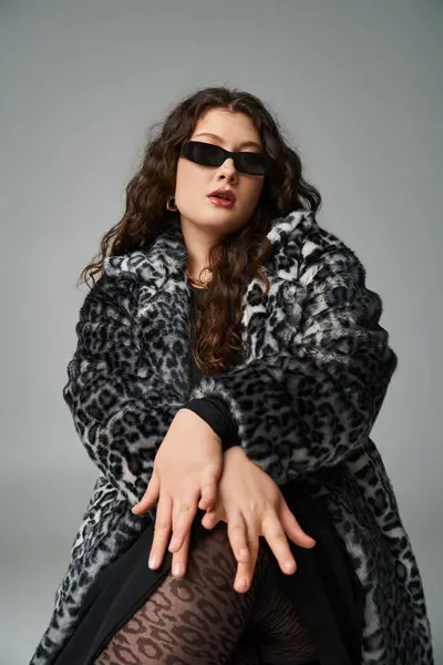 Attractive curvy girl in leopard fur coat and sunglasses sitting with cross legs and hands on knees — Stock Photo