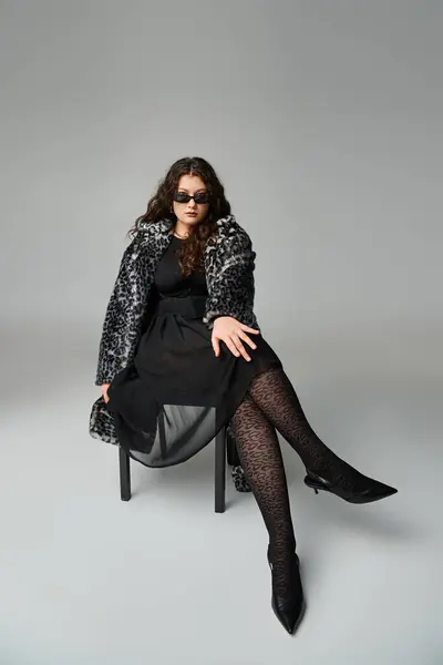 Пишна жінка в шубці леопарда і сонцезахисних окулярах, що сидять на стільці з поперечними ногами і рукою на колінах — стокове фото