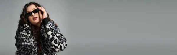 Banner of curvy woman in leopard fur coat touching with hand to sunglasses on grey background — Stock Photo