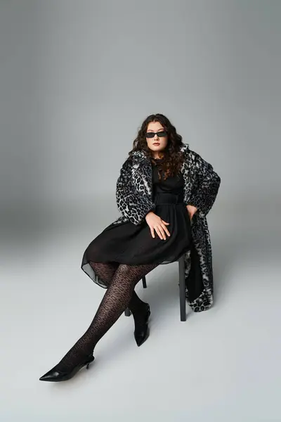 Розслаблений спокусливий плюс розмір жінки в пальто і сонцезахисні окуляри, сидячи на чорному стільці — стокове фото