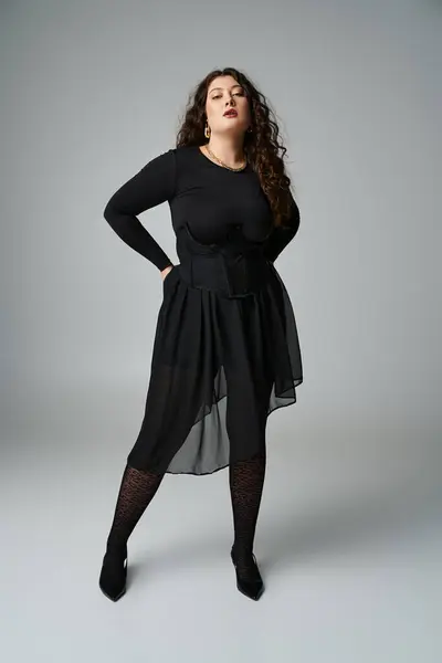Stylish plus size woman in black outfit with hands behind back against grey background — Stock Photo