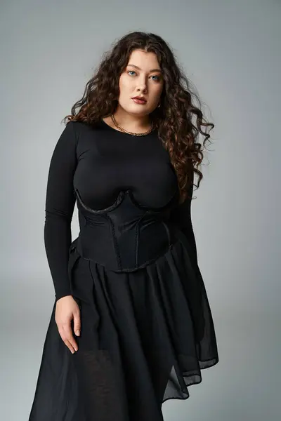 Glamurosa mujer joven de talla grande en traje negro con pelo castaño rizado sobre fondo gris - foto de stock