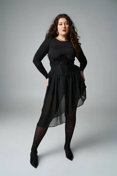 Mulher bonita plus size em roupa preta com cabelo encaracolado e mãos em quadris contra fundo cinza — Fotografia de Stock