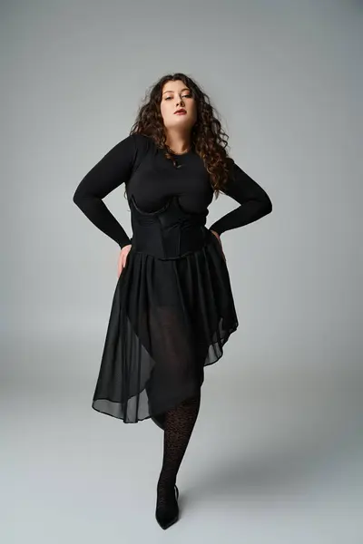 Милий плюс розмір жінки в чорному вбранні позує руками на стегнах і зігнута нога на сірому фоні — стокове фото