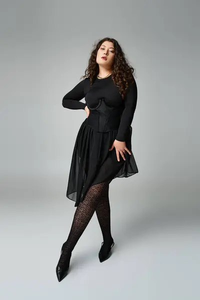Séduisante femme courbée en tenue noire mettant la jambe en avant avec la main à la taille sur fond gris — Photo de stock