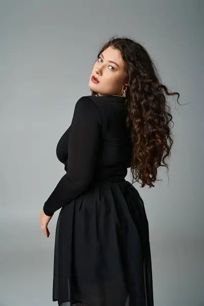 Attractive curvy woman in black outfit looking to camera from behind back on grey background — Stock Photo