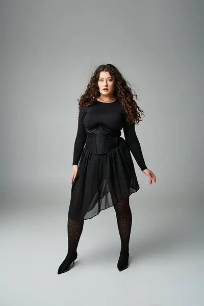 Attractive curvy young girl in black stylish outfit standing with legs wide apart on grey background — Stock Photo
