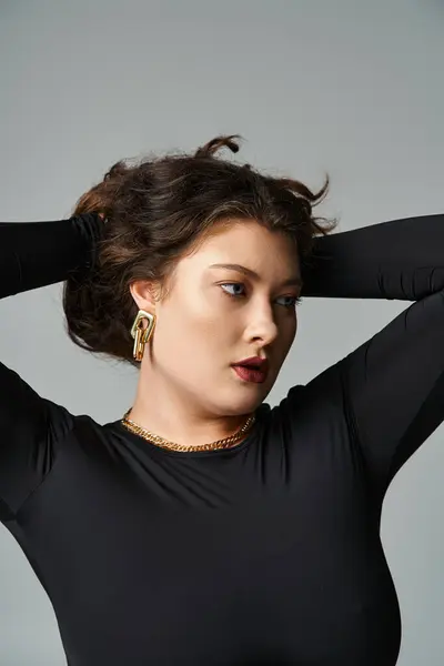 Portrait of pretty plus size woman in her 20s putting hands behind head and holding hair on grey — Stock Photo