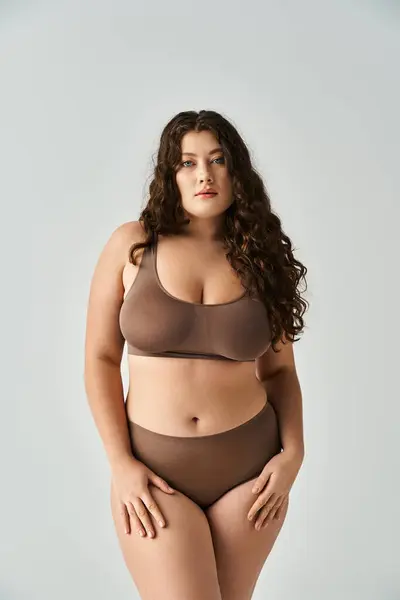 Attractive plus size woman in brown lingerie with curly hair and blue eyes posing on grey background — Stock Photo