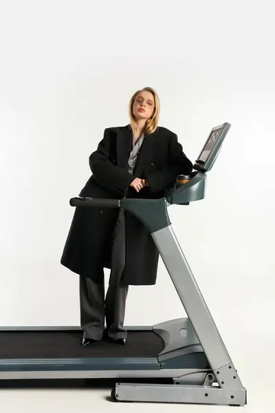 Attractive graceful woman in stylish glasses and coat posing on treadmill and looking at camera — Stock Photo