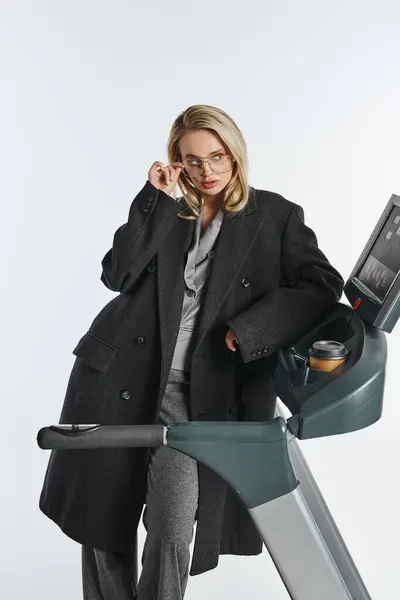 Appealing graceful woman in stylish black coat with trendy glasses posing on treadmill with coffee — Stock Photo
