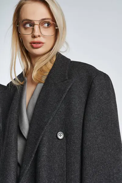 Elegante mujer de moda con el pelo rubio y gafas elegantes en el abrigo negro posando y mirando hacia otro lado - foto de stock