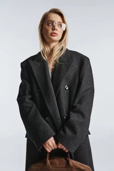 Elegante mujer de moda con el pelo rubio y gafas elegantes en el abrigo negro posando y mirando hacia otro lado - foto de stock