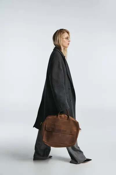 Atractiva mujer elegante con cabello rubio en negro elegante abrigo sosteniendo bolsa marrón y mirando hacia otro lado - foto de stock