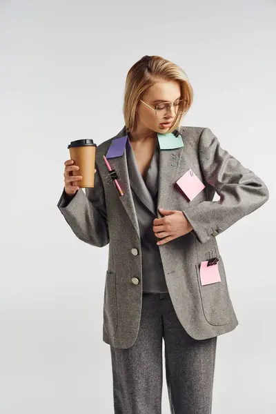 Mulher atraente na moda em terno cinza com artigos de papelaria sobre ele posando com café e olhando para longe — Fotografia de Stock