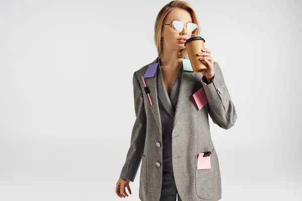 Femme attrayante à la mode en costume gris avec papeterie sur elle posant avec du café et détournant les yeux — Photo de stock
