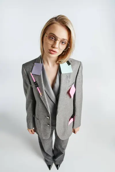 Zierliche Frau mit Brille im grauen Anzug mit Papeterie darauf und Blick in die Kamera vor grauem Hintergrund — Stockfoto
