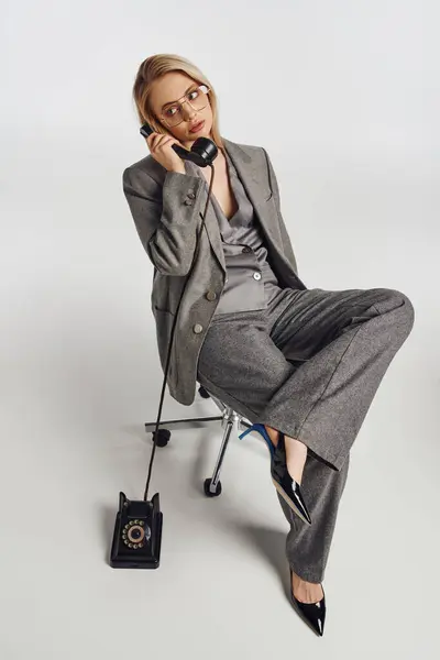 Attraktive schicke Frau mit stylischer Brille im grauen Anzug sitzt auf einem Stuhl und telefoniert mit einem Retro-Handy — Stockfoto