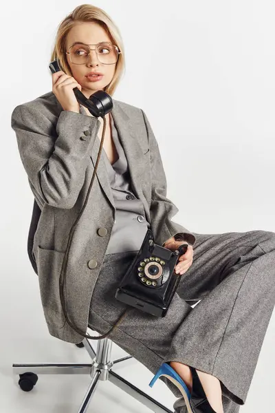 Attraktive schicke Frau mit stylischer Brille im grauen Anzug sitzt auf einem Stuhl und telefoniert mit einem Retro-Handy — Stockfoto