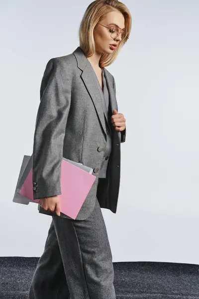 Sofisticada mujer elegante con gafas de estilo en traje gris posando con papeleo y mirando hacia otro lado - foto de stock