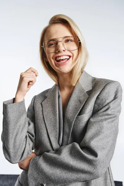Mujer atractiva alegre con gafas en traje elegante gris posando sobre fondo gris y mirando hacia otro lado - foto de stock