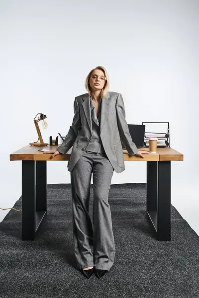 Attraente elegante donna d'affari in abito grigio in posa accanto al tavolo con computer portatile e guardando la fotocamera — Foto stock