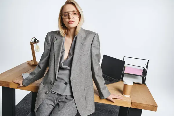Atraente elegante empresária em terno cinza posando ao lado da mesa com laptop e olhando para a câmera — Fotografia de Stock