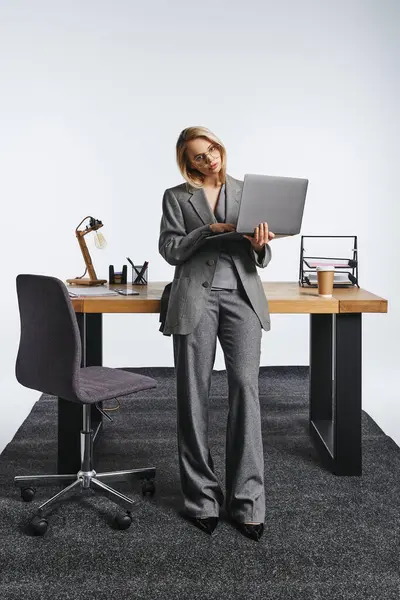 Appealing elegant businesswoman in gray suit posing next to table with laptop and looking at camera — Stock Photo