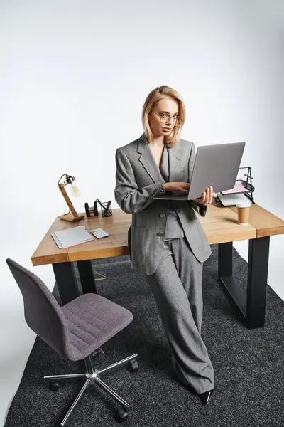 Привлекательная элегантная деловая женщина в сером костюме позирует рядом со столом с ноутбуком и смотрит в камеру — стоковое фото