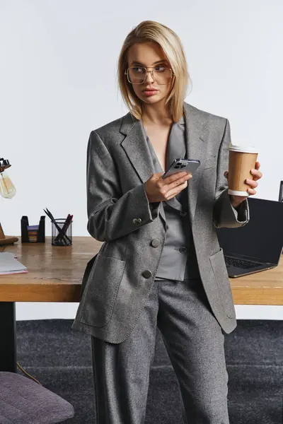 Fashionable young businesswoman in gray stylish suit holding smartphone and coffee and looking away — Stock Photo