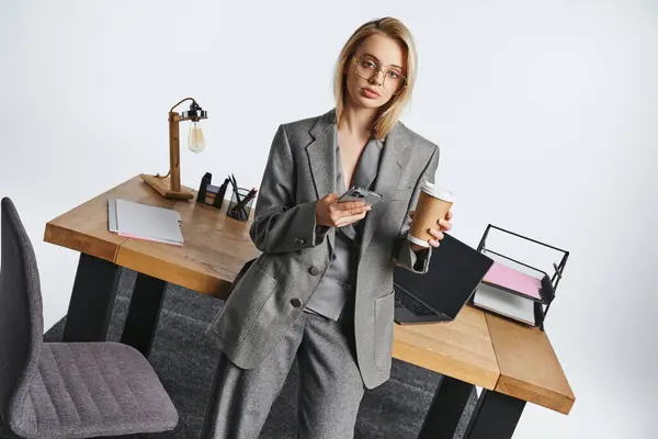 Die elegante Geschäftsfrau im grauen schicken Anzug hält Smartphone und Kaffee in der Hand und blickt in die Kamera — Stockfoto