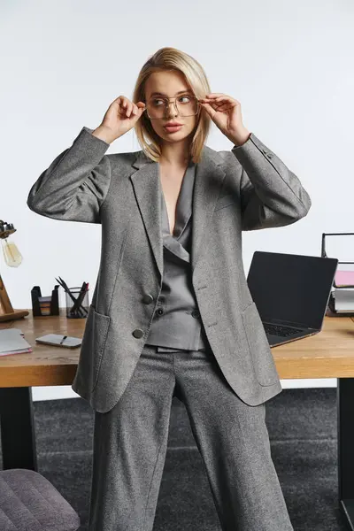 Hermosa mujer de negocios elegante en traje inteligente gris posando junto a la mesa en la oficina y mirando hacia otro lado - foto de stock
