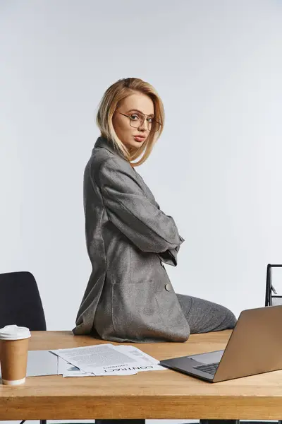 Gut aussehende elegante Geschäftsfrau im grauen schicken Anzug sitzt auf dem Tisch und schaut in die Kamera — Stockfoto