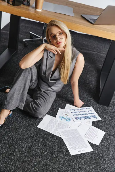 Debonair mulher loira em terno cinza elegante trabalhando no chão com seus papéis e olhando para a câmera — Fotografia de Stock