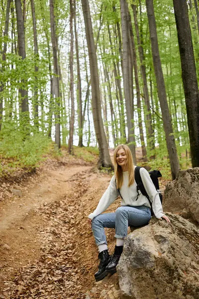 Smiling blonde female hiker wearing backpack, resting on rocks, enjoying the forest view — Stock Photo
