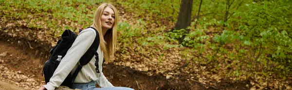 Entspannt lächelnde blonde Wanderin in Pullover und Rucksack auf einem Felsen im Wald sitzend, Banner — Stockfoto
