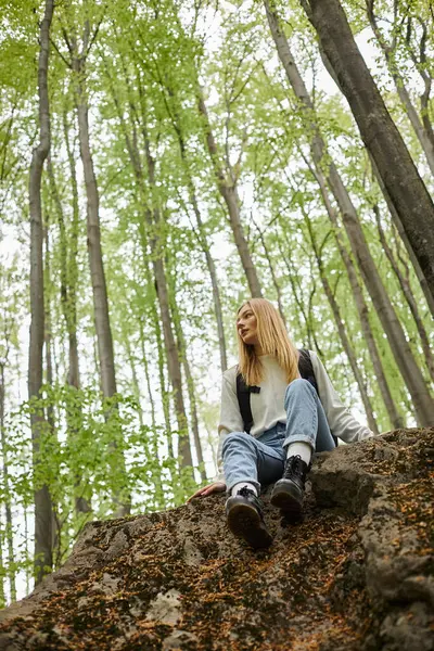 Calm relaxed blond girl hiking in forest sitting on rock and wearing sweater and backpack — Stock Photo