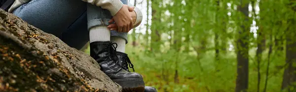 Foto recortada de la mujer excursionista abrazando sus piernas con jeans y botas de senderismo en el bosque, pancarta - foto de stock