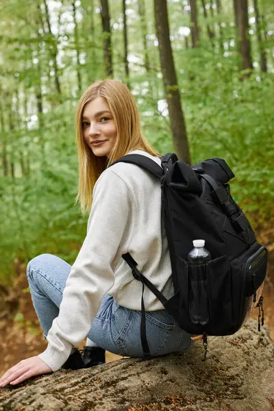 Back view portrait of carefree blonde hiker in cozy, sweater sitting in forest and looking at camera — Stock Photo