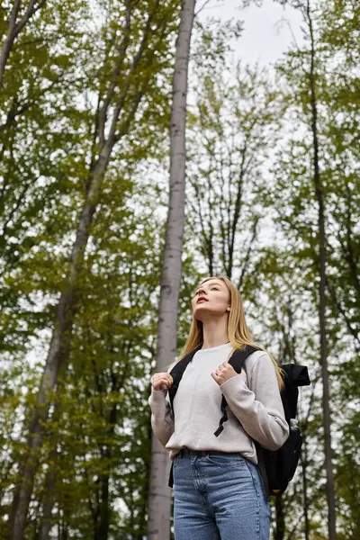 Inspired blonde female trekker holding her backpack, wearing sweater, looking through the woods — Stock Photo