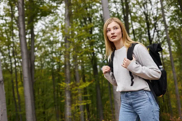 Curieuse femme blonde tenant son sac à dos, portant un pull et un jean, regardant à travers les bois — Photo de stock