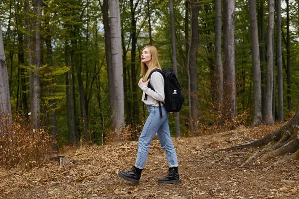 Donna bionda avventuriera in accogliente, maglione e jeans, passeggiando nel bosco con curiosità — Foto stock