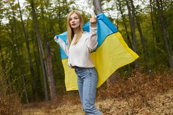 Mujer rubia fuerte y orgullosa de pie en el bosque con la bandera de Ucrania, patriótica - foto de stock