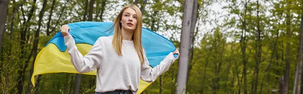 Supportive independent blonde woman standing in the forest with the Ukrainian flag waving it, banner — Stock Photo