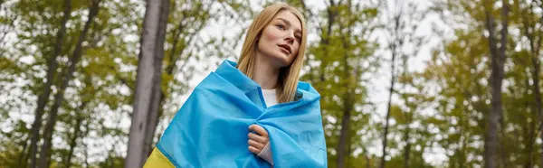 Patriotic woman standing in forest covering body with Ukrainian flag and looking away, banner — Stock Photo