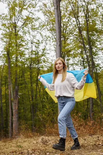 Mujer rubia bonita de pie en el bosque cubriendo el cuerpo con la bandera nacional de Ucrania como capa - foto de stock