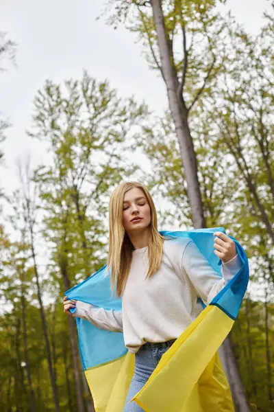 Calma mujer rubia al aire libre en el bosque que cubre el cuerpo con la bandera nacional de Ucrania como capa - foto de stock