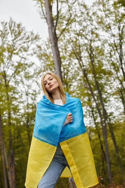 Ernstes blondes Mädchen, das im Wald die ukrainische Flagge über ihrem Körper hält und in die Kamera blickt — Stockfoto